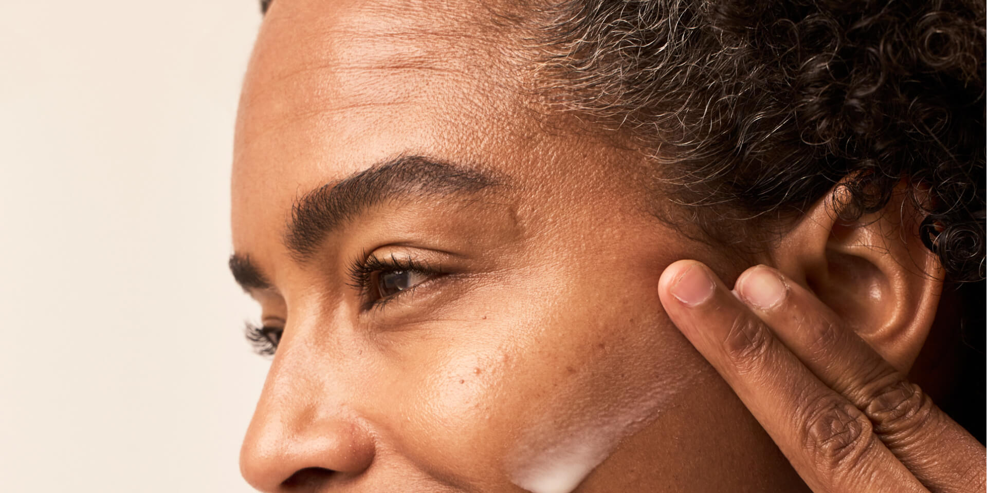 Side view of a face with fingers touching the cheek, showing a small amount of cream on the skin under soft lighting
