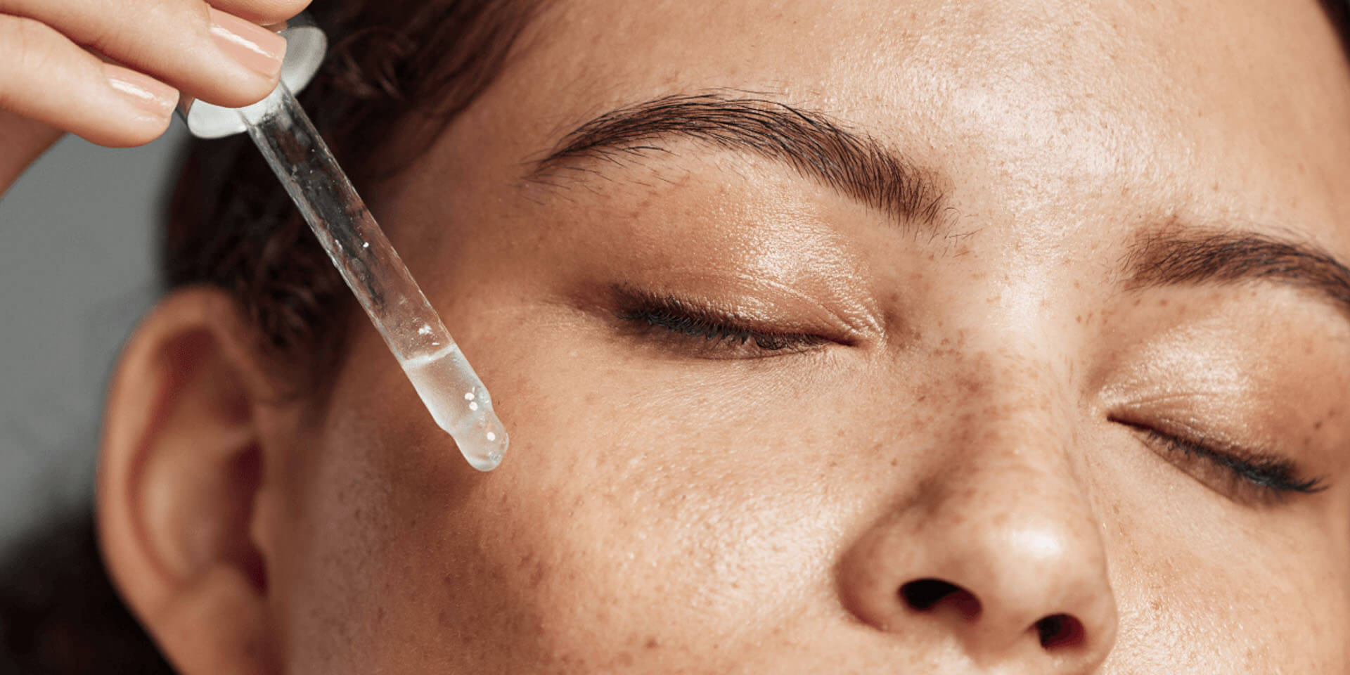 Close-up of woman with eyes closed applying clear serum to her temple using a glass dropper