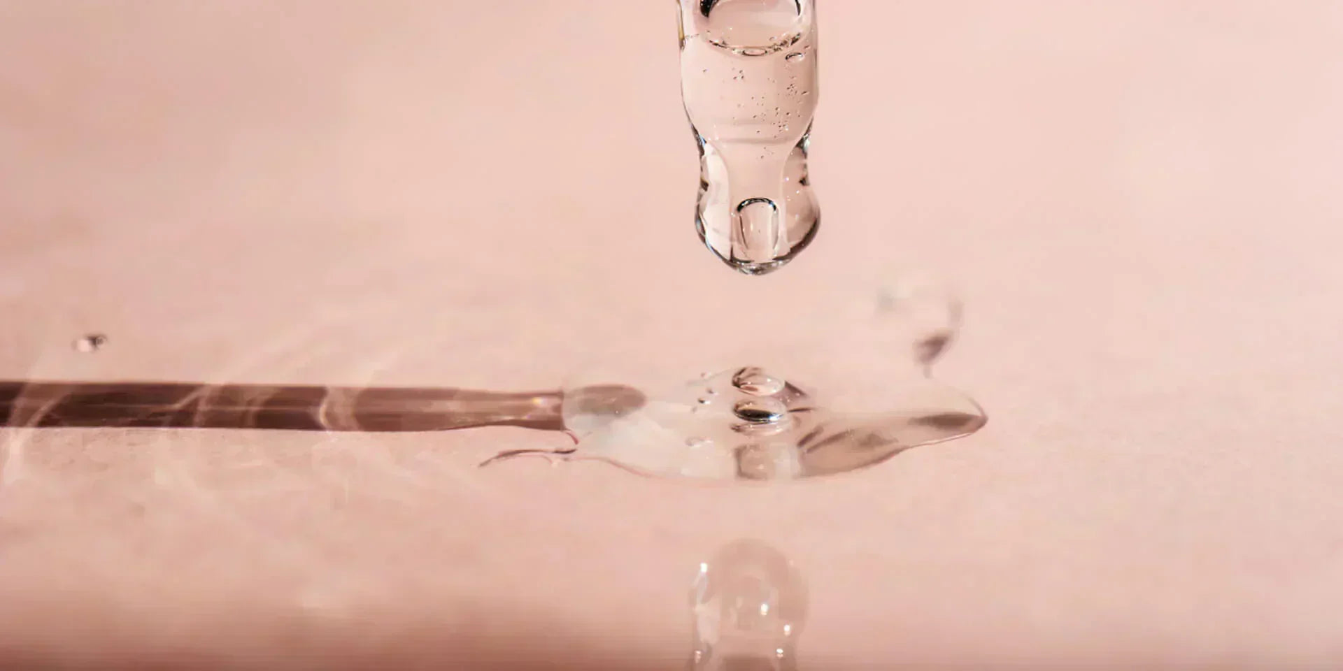 Macro view of a clear liquid drop suspended above a small pool on a smooth, light‑colored surface