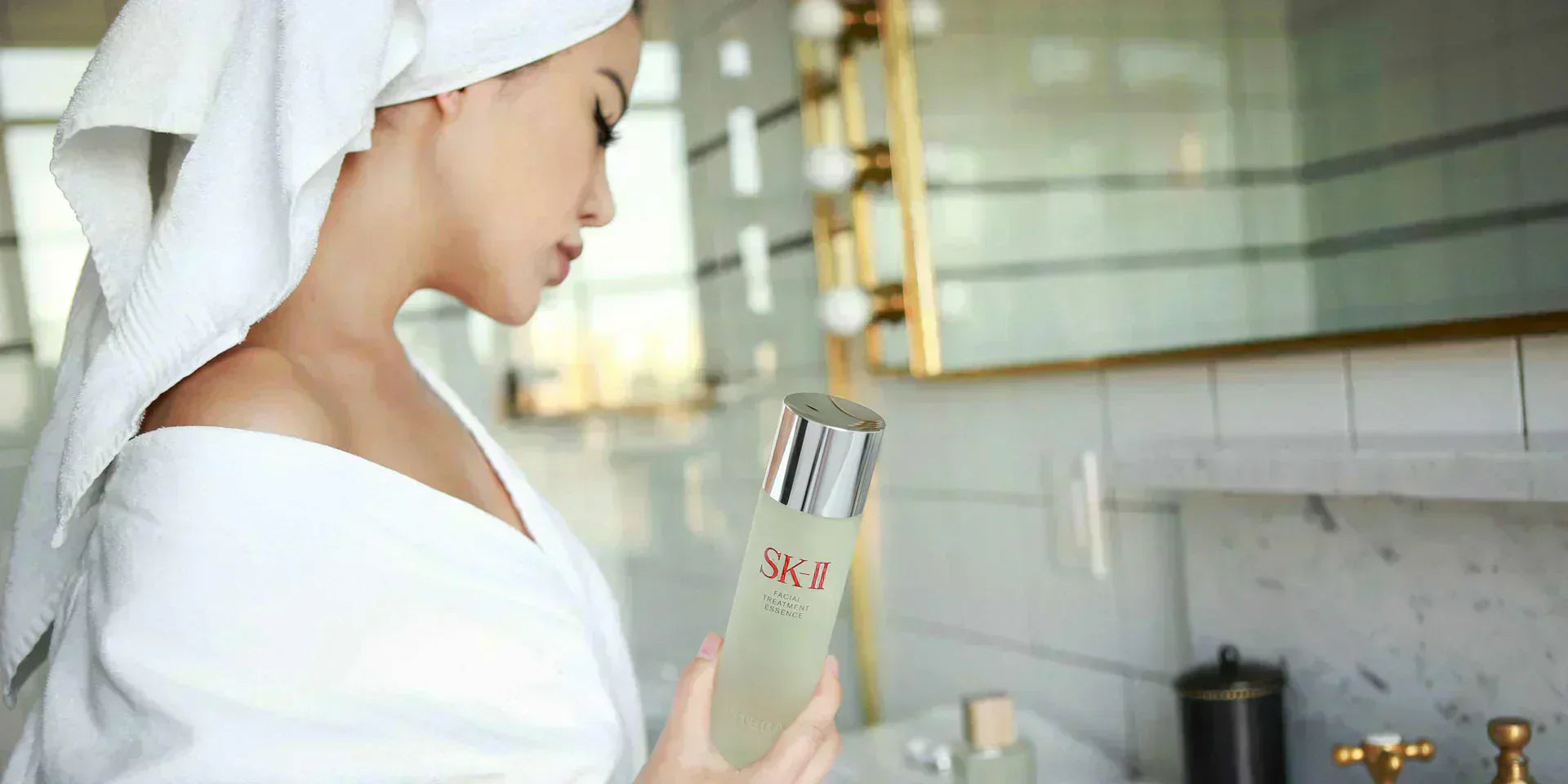 Woman with towel wrapped around head holding SK-II Facial Treatment Essence bottle in a bathroom setting with mirror and sink