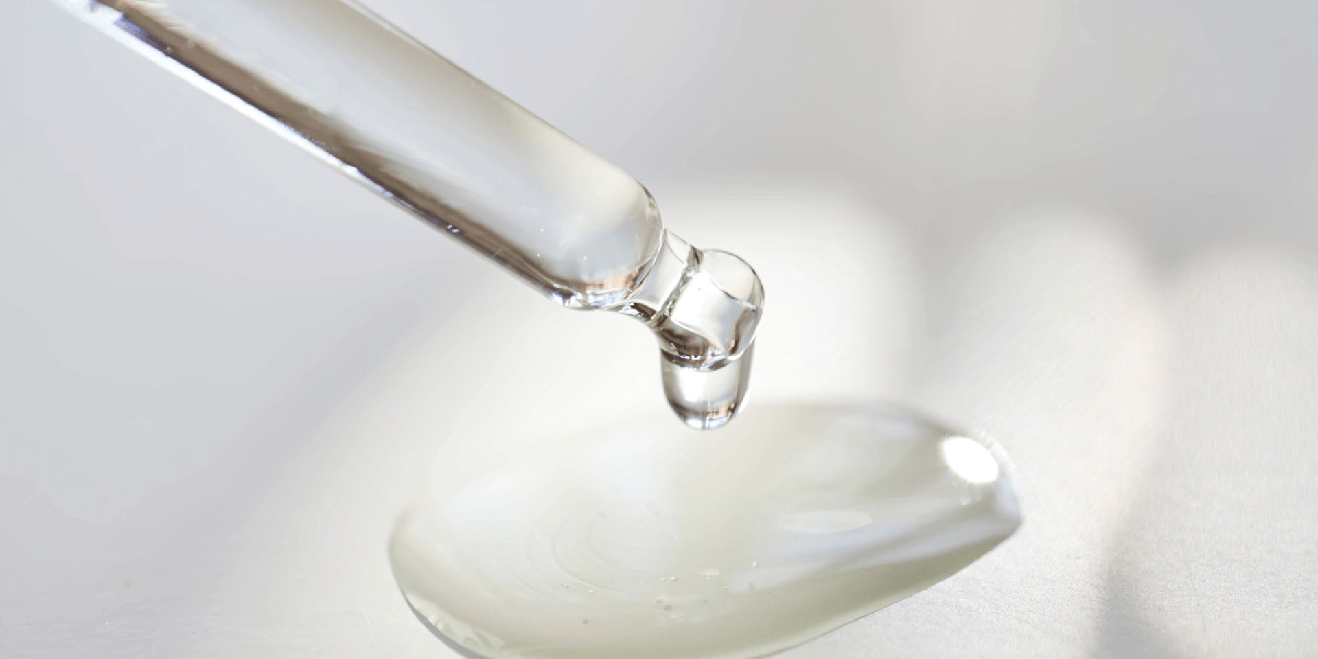 Close-up of a glass dropper releasing a clear liquid droplet onto a smooth surface, forming a small glossy pool