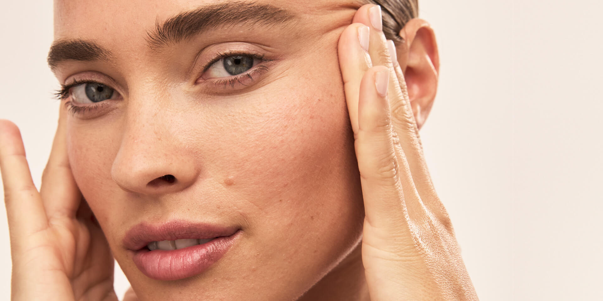 Close-up of face with hands placed along cheeks, showing skin texture and natural lines around the eye area on a light background