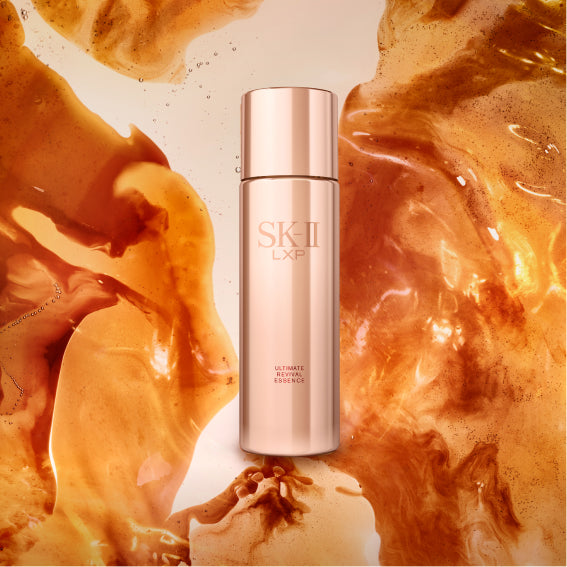 SK-II LXP Ultimate Revival Essence bottle in metallic gold packaging surrounded by flowing amber liquid on a light background