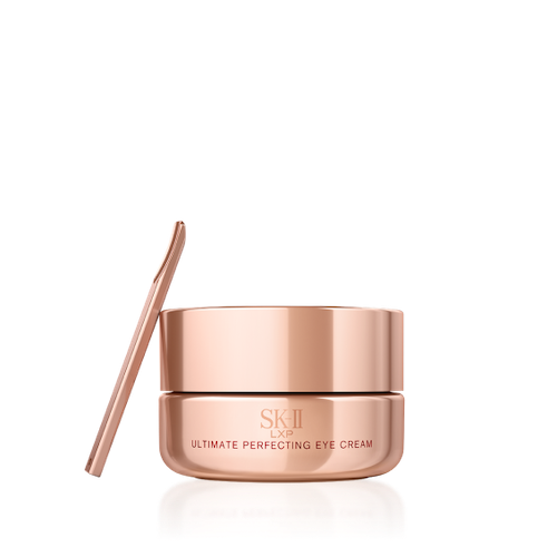 SK-II LXP Ultimate Revival Eye Cream jar with metallic gold finish and spatula placed beside it, surrounded by flowing amber liquid